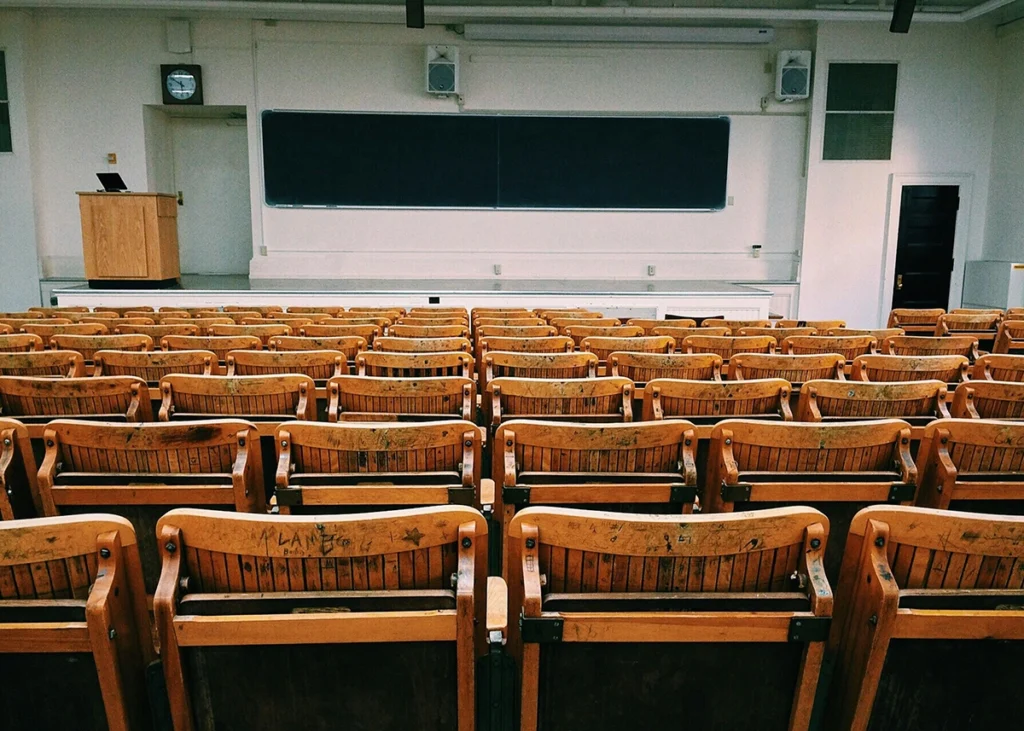 Aula vacía con filas de asientos de madera frente a una pizarra y un atril.