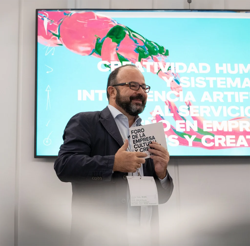 Juan Pastor Bustamante hablando en el Foro de la Empresa Cultural y Creativa, sosteniendo un cuaderno frente a una pantalla de presentación.